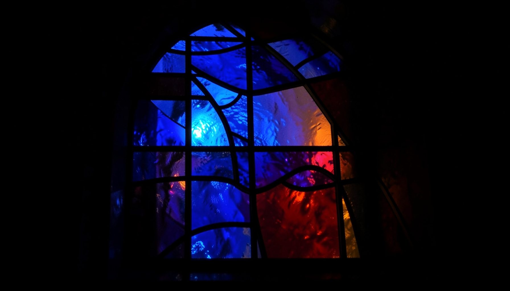 A stained-glass composition with bold leaded segments in deep indigo, amber, and ruby tones, casting warm filtered light across a dark interior space