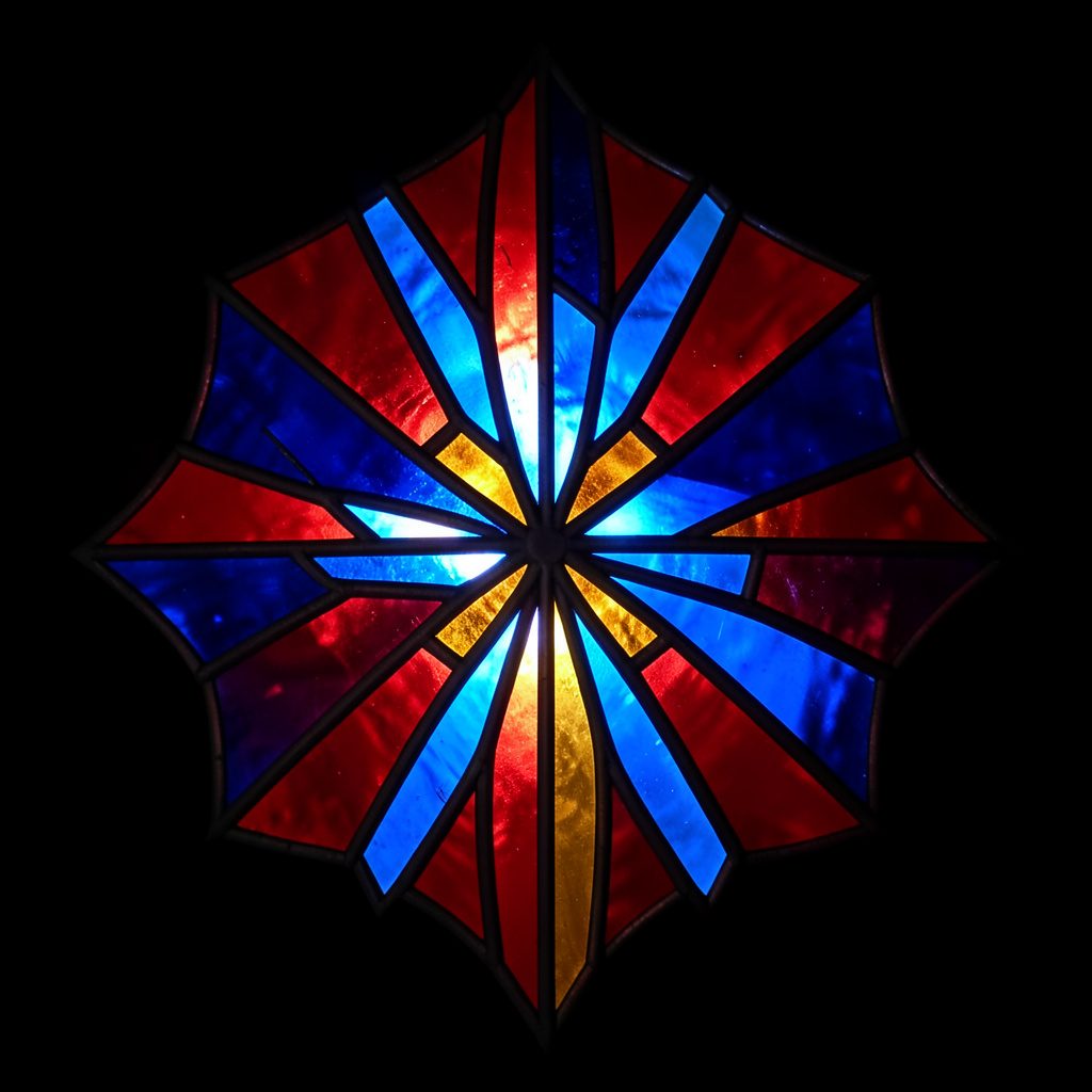 Stained-glass emblem with bold leaded segments in deep red, cobalt blue, and amber, forming a radiant geometric burst against a dark ground
