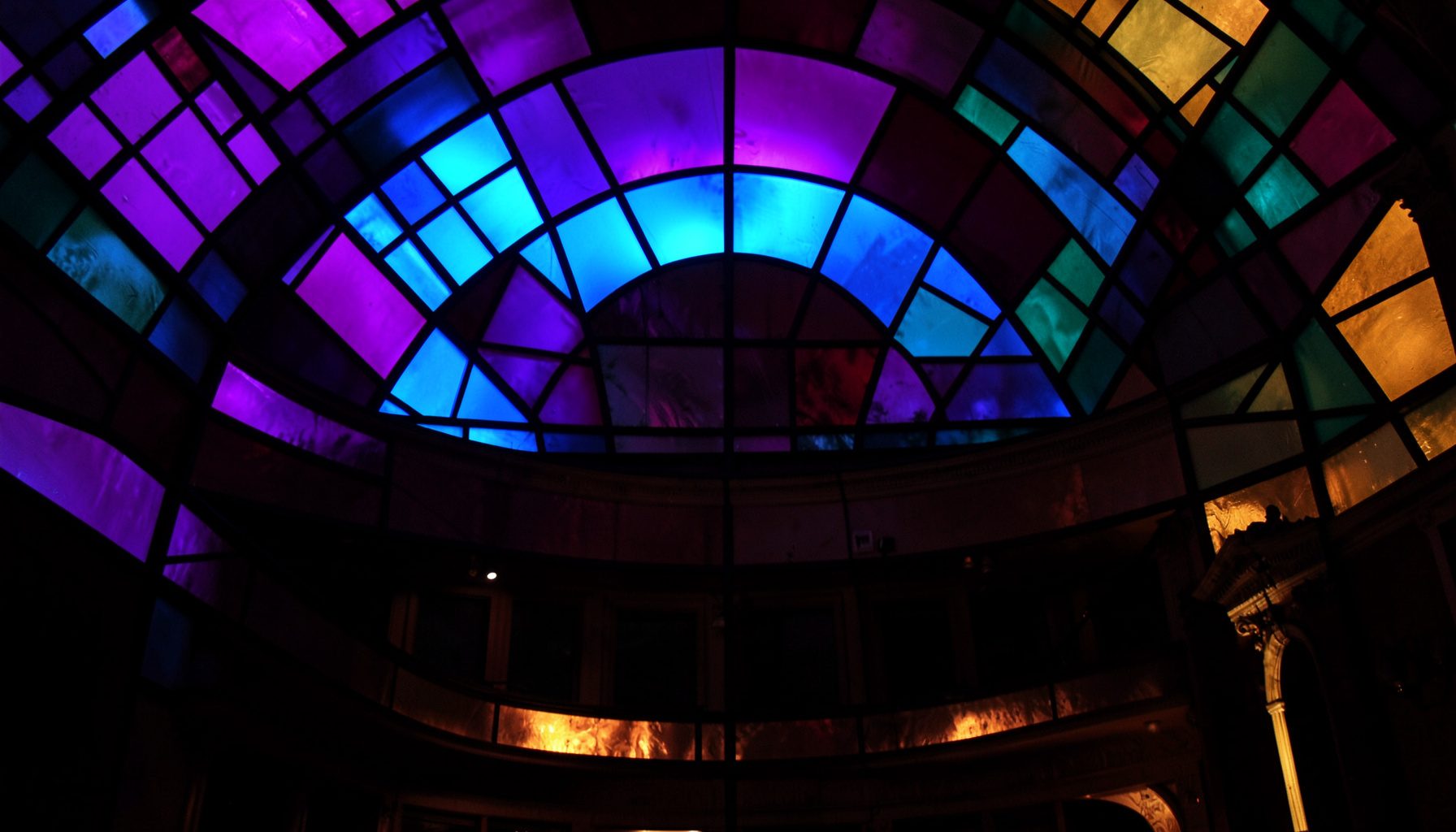 Stained-glass composition with bold leaded segments in deep violet, teal, and warm amber, arranged in layered geometric arcs suggesting a theatre interior